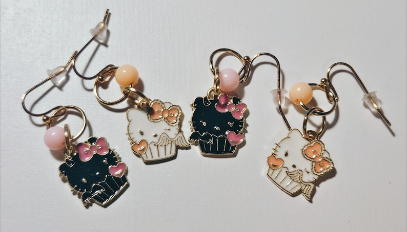 May include: Four gold-toned dangle earrings. Each earring features a charm shaped like a cupcake with a Hello Kitty design. The charms are black and white with pink accents. Each earring has a pink and peach-colored bead.