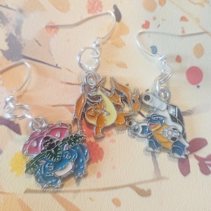 Puede incluir: Un par de pendientes de plata con abalorios de esmalte de colores que representan personajes de Pokemon. Los abalorios son de Bulbasaur, Charmander y Squirtle.