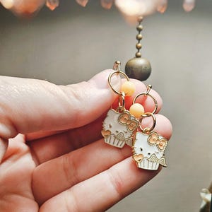 May include: Gold-toned hoop earrings with white enamel cupcake charms featuring a bow and heart details. Each earring has a small peach-colored bead. The earrings are held in a hand.