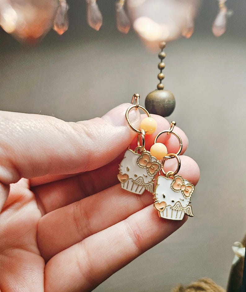 May include: Gold-toned hoop earrings with white enamel cupcake charms featuring a bow and heart details. Each earring has a small peach-colored bead. The earrings are held in a hand.