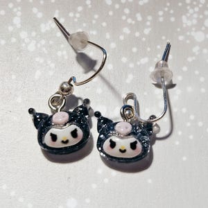 Pode incluir: Um par de brincos pendurados com um design de personagem de desenho animado. Os brincos têm um acabamento preto e brilhante com um rosto branco, detalhes em rosa e ganchos de orelha prateados. Os brincos são projetados para serem usados por qualquer pessoa.