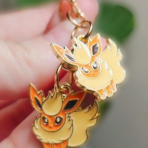 Puede incluir: Llaveros de metal dorado con dos personajes de dibujos animados Flareon. Las figuras de Flareon tienen pelaje naranja y amarillo con detalles en negro y están unidas a una cadena dorada. El fondo está borroso.
