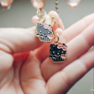 May include: A pair of dangle earrings featuring a black and gray cartoon cat design with a pink bow and heart accents. The earrings are suspended from gold-colored hoops.