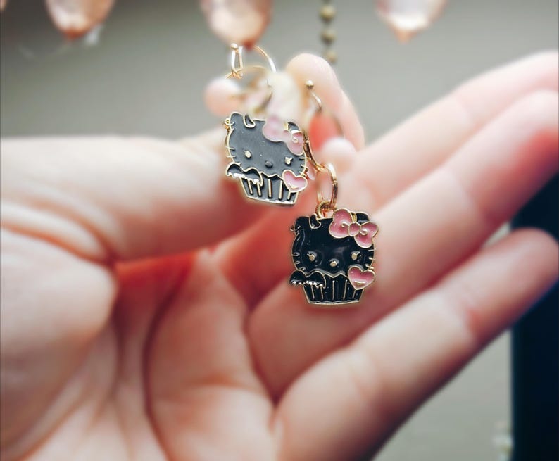 May include: A pair of dangle earrings featuring a black and gray cartoon cat design with a pink bow and heart accents. The earrings are suspended from gold-colored hoops.