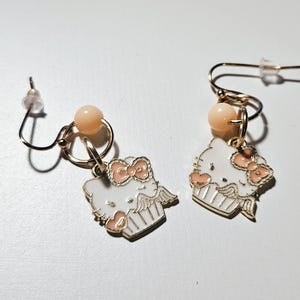 May include: A pair of gold-colored earrings featuring a white cartoon character design with a pink bow and accents. Each earring has a small peach-colored bead and a gold-colored hook.