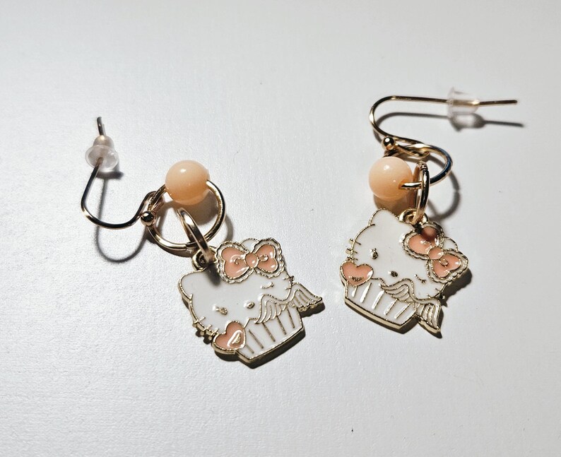 May include: A pair of gold-colored earrings featuring a white cartoon character design with a pink bow and accents. Each earring has a small peach-colored bead and a gold-colored hook.