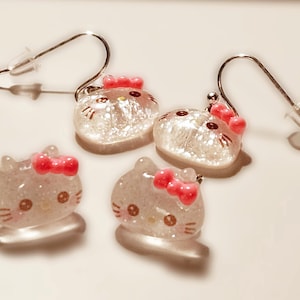 Pode incluir: Brincos Hello Kitty transparentes e brilhantes com laços cor-de-rosa. Os brincos têm ganchos prateados e tampões de plástico transparente. Dois pares de brincos são visíveis, com os rostos de Hello Kitty virados em direções diferentes.