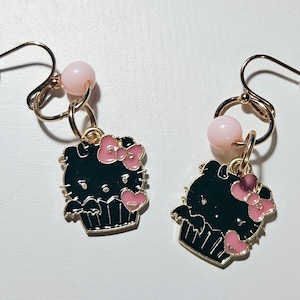 May include: A pair of dangle earrings featuring a black cupcake design with pink bows and hearts. Each earring has a gold-colored hook, a pink bead, and a clear earring back. The earrings are set against a white background.
