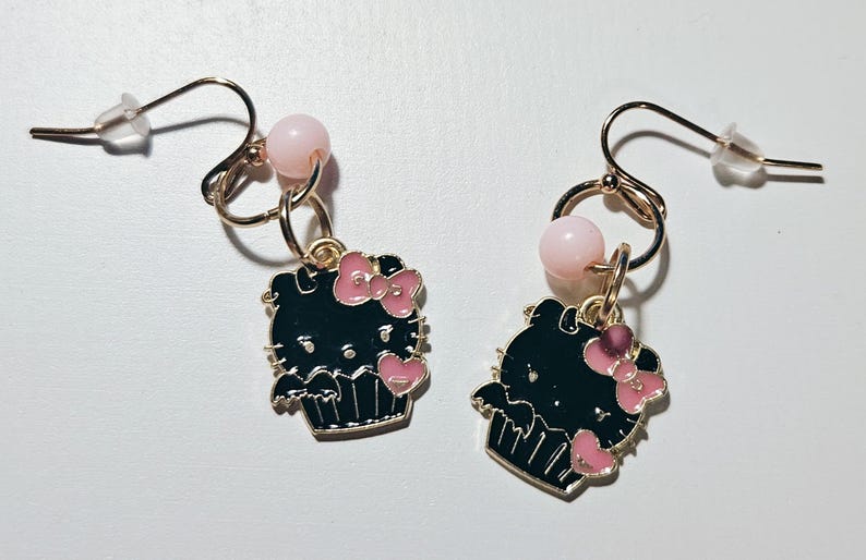 May include: A pair of dangle earrings featuring a black cupcake design with pink bows and hearts. Each earring has a gold-colored hook, a pink bead, and a clear earring back. The earrings are set against a white background.