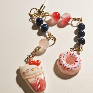 May include: A charm featuring a Hello Kitty-themed drink and a pink donut with sprinkles. The charm is connected by a chain of blue, pink, and red beads. The charm has a gold-coloured clasp and a white string.