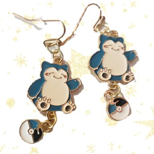 May include: A pair of gold-colored dangle earrings. Each earring features a blue and white cartoon character with a smiling face and a small, round charm with black, white, and blue accents.