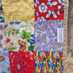 May include: A patchwork quilt with a variety of colorful fabrics, including floral prints, butterflies, and geometric patterns. The quilt is made with a variety of fabrics, including cotton and linen. The quilt is edged with a tan fabric and has a label that reads "Second Sanctuary Studio" and "www.secondsanctuary.etsy.com".