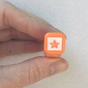 May include: An orange, rectangular stamp featuring a white square and a star design. The stamp is held in a hand against a white background. The stamp is a crafting tool.