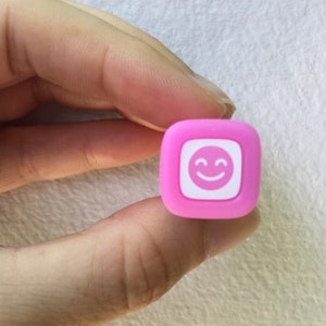 May include: A small pink square stamp with a white smiley face design.