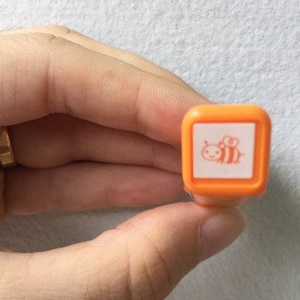 May include: Orange rubber stamp with a white bee design. The bee is facing left and has a small smile.