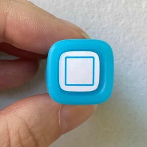 May include: A small, turquoise square with rounded corners and a white square design in the centre. The object is held in a hand against a white background. It resembles a button or a small decorative element.