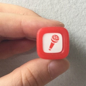 May include: A red square-shaped stamp with a white microphone icon inside. The stamp is being held by a hand.