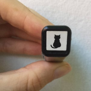 May include: Black and white stamp with a silhouette of a cat sitting on a white background. The stamp is in a black square frame.