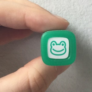 May include: Green plastic stamp with a white frog design. The frog has a smiling face.
