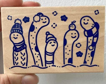May include: A wooden stamp with a blue design of five friendly worm characters wearing winter hats and scarves. The worms are standing in a row on a snowy background with snowflakes.
