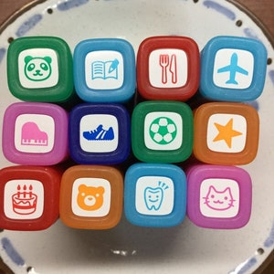 May include: A set of 12 colourful square rubber stamps with various images, including a panda, a book, a fork and knife, an aeroplane, a piano, a trainer, a football, a star, a birthday cake, a bear, a tooth, and a cat.