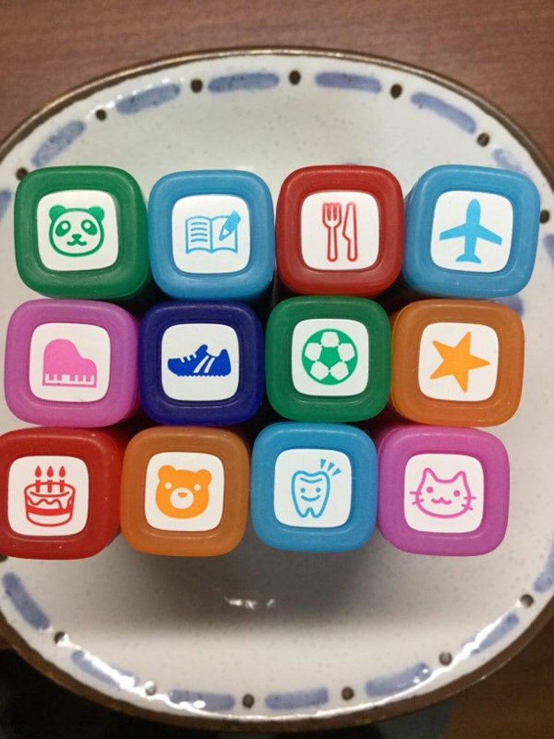 May include: A set of 12 colourful square rubber stamps with various images, including a panda, a book, a fork and knife, an aeroplane, a piano, a trainer, a football, a star, a birthday cake, a bear, a tooth, and a cat.