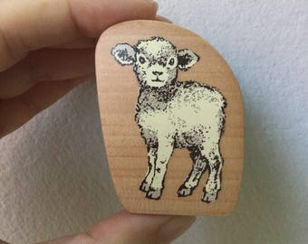 May include: A wooden stamp with a black and white illustration of a lamb. The lamb is standing on all four legs and has a white face with black eyes and a black nose.