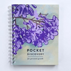 May include: A purple and white floral design on the cover of a spiral bound notebook. The notebook is titled "Pocket Discovery" for personal growth.