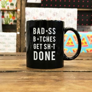 Könnte beinhalten: Schwarze Keramik-Kaffeetasse mit weißem Text, der "BAD*SS B*TCHES GET SH*T DONE" lautet.