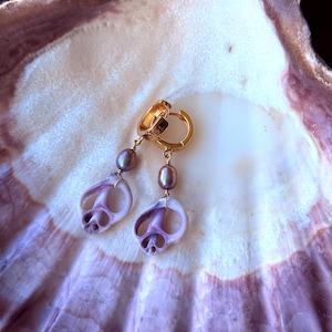 May include: Gold hoop earrings with dangling purple shell-shaped charms and purple pearl accents. The earrings are displayed on a large, light purple and white seashell. The earrings have a unique, beachy aesthetic.