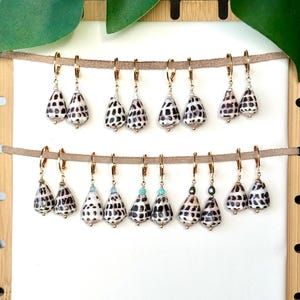 May include: A collection of seashell earrings with gold-colored hoops. The earrings feature a teardrop-shaped shell design with black and white patterns. Some earrings have small, colorful beads. The earrings are displayed on a neutral background.