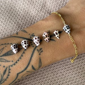May include: A gold chain bracelet with six small, cone-shaped seashells. The shells are white with black and brown spots. The bracelet is on a person's arm, with a tattoo visible.