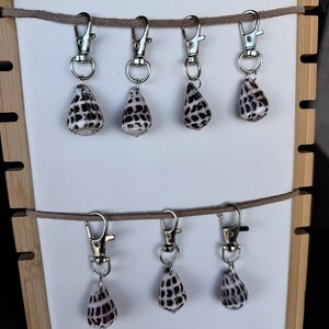 May include: A display of keychains featuring teardrop-shaped seashells with black and white patterns. Each shell is attached to a silver-colored clasp. The keychains are suspended from a brown cord against a white background.