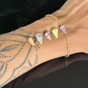May include: A gold chain bracelet featuring six small, colorful seashells. The seashells are in shades of orange, silver, red, yellow, and purple. The bracelet is worn on a wrist with a tattoo.