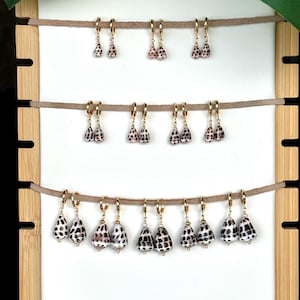 May include: Gold hoop earrings with teardrop-shaped pendants made from natural seashells. The pendants have a speckled pattern of black and brown spots on a cream background. The earrings are displayed on a white background.