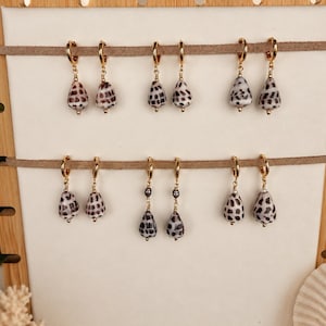 May include: Five pairs of gold hoop earrings featuring dangling teardrop-shaped seashell charms with brown and black spotted patterns. The earrings are displayed on two horizontal tan suede strips attached to a light-colored rectangular board.