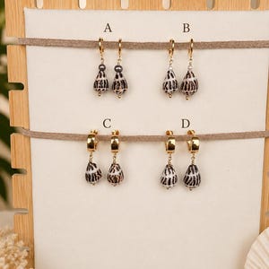May include: Four pairs of drop earrings labeled A, B, C, and D. Pairs A and B feature small gold hoops with dark grey beads and patterned shell charms. Pairs C and D feature thicker gold hoops with patterned shell charms hanging from gold chains.