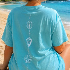 May include: A light blue t-shirt featuring a white seashell design. The design includes three different seashell illustrations connected by a thin white line. The shirt is short-sleeved and made of a soft material.