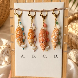 May include: Four gold-toned lobster clasp keychains labeled A, B, C, and D, each featuring a spiral orange and white seashell, small dangling beads in blue, purple, pink, or green, and a tiny white shell charm hanging from a gold chain.