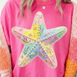 Puede incluir: Una camiseta rosa brillante con una gran estrella de mar decorativa. La estrella de mar está hecha de parches de tela coloridos con estampados florales y está delineada con pequeñas perlas blancas. Se lleva un cárdigan de punto grueso multicolor sobre la camisa.