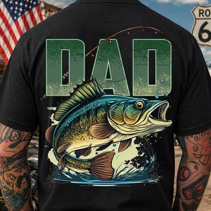 Pu&ograve; includere: T-shirt nera con un'illustrazione verde e blu di un pesce che salta fuori dall'acqua, con la parola "DAD" in lettere verdi sopra. Il design include una lenza. Lo sfondo mostra un cartello della Route 66 e una bandiera americana.