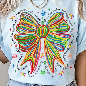 May include: Light blue t-shirt featuring a large, colorful bow design. The bow is made of multiple lines in various colors. The shirt also has text around the bow that says "brave, creative, friendly, strong, love, kind, enough, worthy".