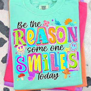 Könnte beinhalten: Hellblaues Comfort Colors T-Shirt mit dem farbenfrohen Schriftzug "Be the REASON some one SMILES Today". Das Design umfasst Blumen, Schmetterlinge und ein Smiley. Das T-Shirt liegt gefaltet auf anderen bunten Shirts.