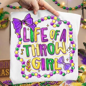 May include: White tote bag with the text "LIFE OF A THROW GIRL" in green, purple, and yellow. The design includes a purple bow, musical notes, and a beaded necklace border. The bag is held up by a person's hand.