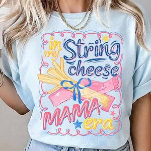 May include: Light blue t-shirt with a colorful graphic. The graphic features the text "in my String cheese MAMA era" with illustrations of string cheese and a bow. The design is outlined in pink and blue.