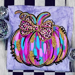 May include: A lavender t-shirt featuring a colorful pumpkin design. The pumpkin has a pink, blue, and purple color scheme with gold glitter accents and a leopard print bow. Black outlines define the pumpkin's shape.