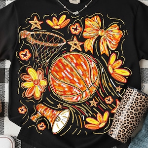May include: Black t-shirt with a basketball-themed graphic. The design includes a basketball, flowers, stars, a bow, and a megaphone, all in orange, yellow, and gold. A leopard-print tumbler is visible on the right.