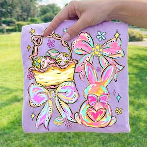 May include: A lilac t-shirt with a vibrant Easter-themed design. The design features a basket of decorated eggs, a bunny with a heart, and bows and flowers. The artwork is outlined in black and has a watercolor effect.