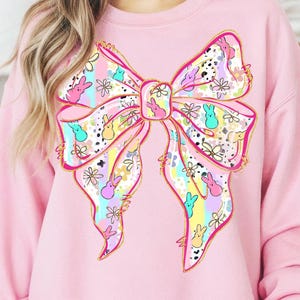 May include: Pink sweatshirt featuring a large, colorful bow design. The bow is decorated with pastel colors, flowers, and cartoon bunny figures. The bow has a gold outline.