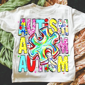 May include: White t-shirt with the word "AUTISM" in colorful, block letters. The letters are filled with puzzle pieces, hearts, and other designs. A large, multi-colored puzzle piece is in the center of the shirt.
